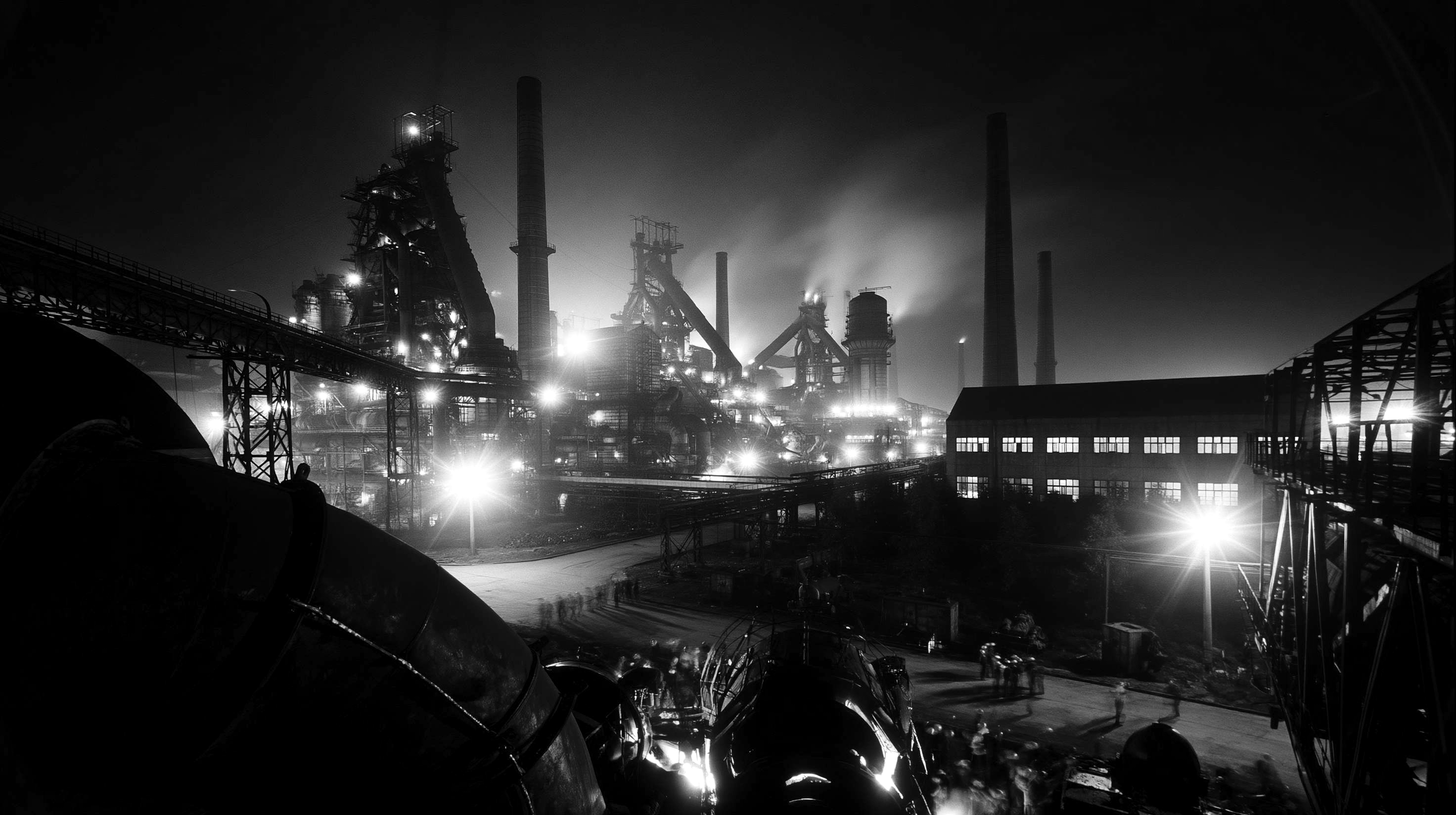 A large industrial plant lit up at night, with workers on the ground and blast furnaces in the distance