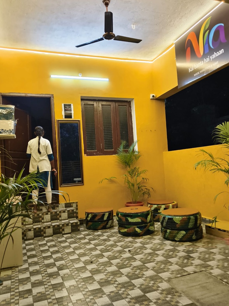 A Nia residence courtyard at dusk with warm yellow walls, tiled flooring, seating and an illuminated Nia sign, as a resident walks toward the doorway